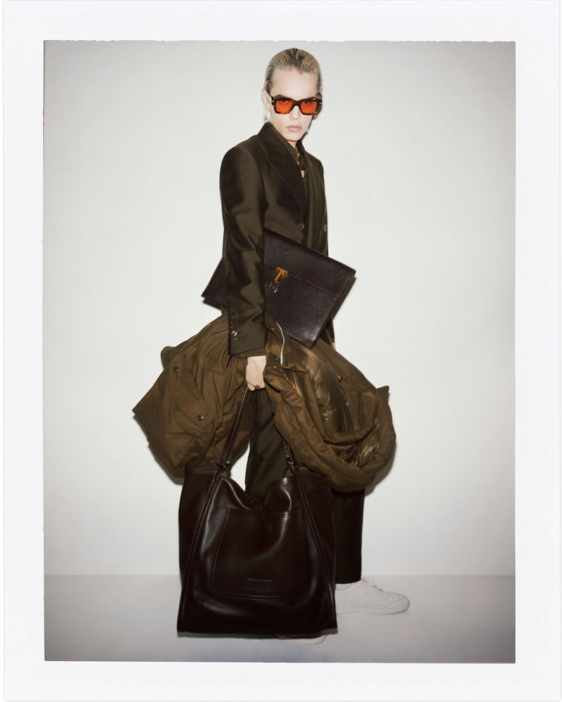 A stylish person with fair hair and sunglasses carries a coat and leather bag against a plain background in a full-length fashion shot.