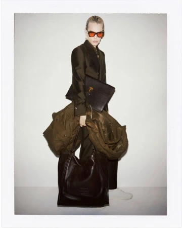 A stylish person with fair hair and sunglasses carries a coat and leather bag against a plain background in a full-length fashion shot.