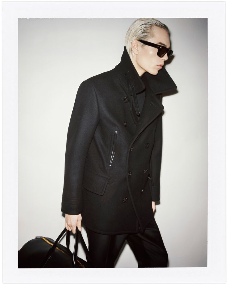 Man with slicked-back hair, sunglasses, and a black coat holds a duffel bag in a fashion studio portrait against a bright background.
