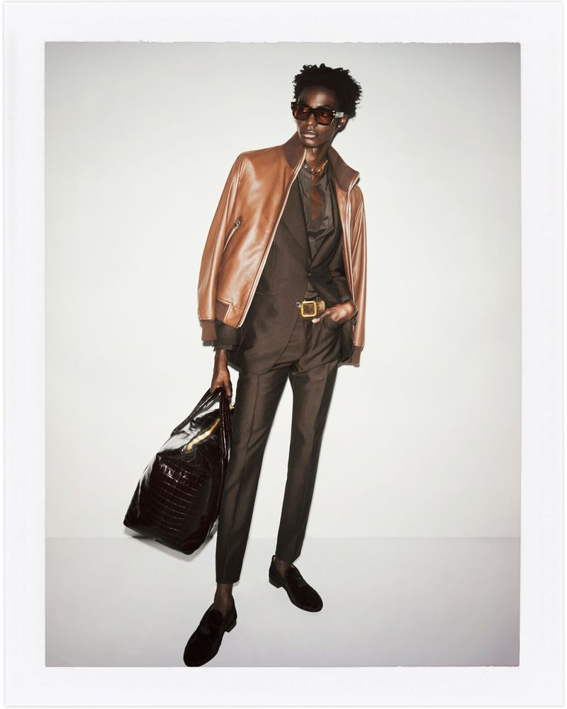 A male model poses in a brown suit and leather jacket, holding a black bag against a plain background in a fashion editorial style photograph.