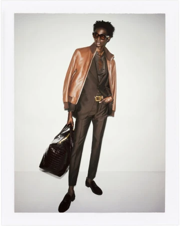 A male model poses in a brown suit and leather jacket, holding a black bag against a plain background in a fashion editorial style photograph.