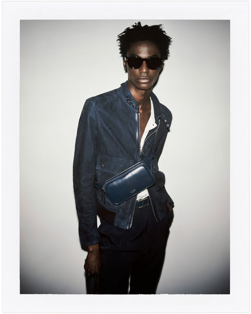 A stylish Black male model in sunglasses and a blue jacket poses against a neutral background, showcasing a sleek, modern fashion look.