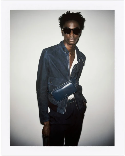 A stylish Black male model in sunglasses and a blue jacket poses against a neutral background, showcasing a sleek, modern fashion look.