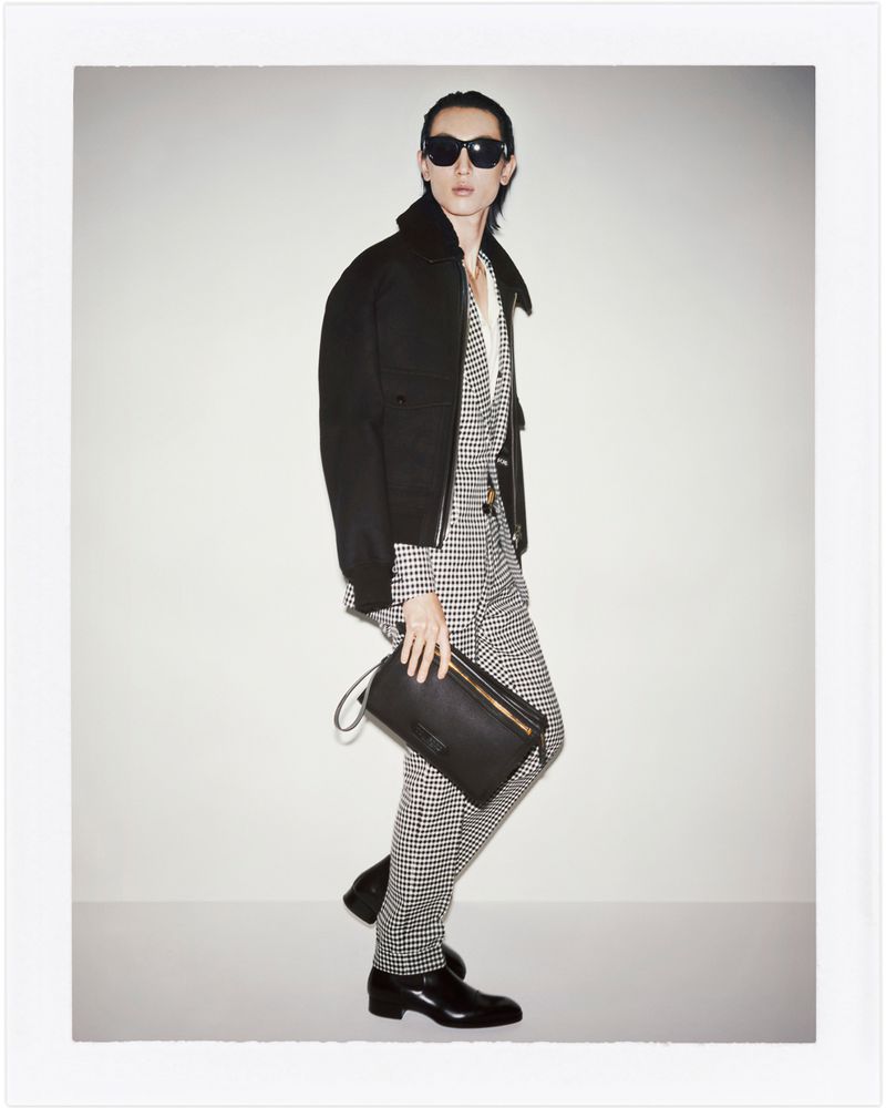 Man in black jacket and houndstooth suit holds a black clutch, posing indoors against a white wall. Fashion shoot with sunglasses.