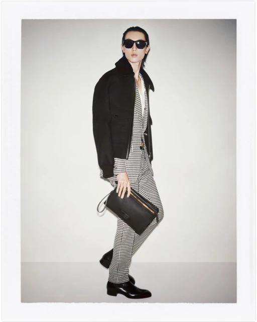 Man in black jacket and houndstooth suit holds a black clutch, posing indoors against a white wall. Fashion shoot with sunglasses.