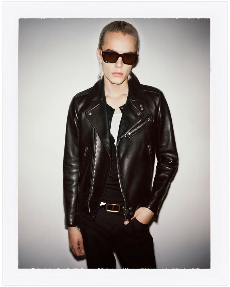 A fashionable adult male model in sunglasses and a black leather jacket poses indoors against a neutral background, looking at the camera.