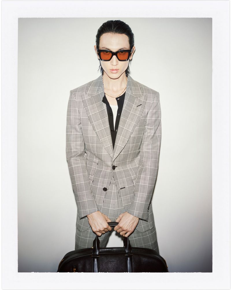 Adult male posing in grey plaid suit, black shirt, and orange sunglasses, holding a leather duffel bag against a white backdrop.