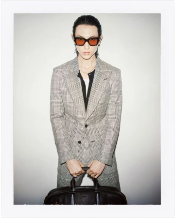 Adult male posing in grey plaid suit, black shirt, and orange sunglasses, holding a leather duffel bag against a white backdrop.