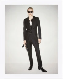 Model in a black suit and sunglasses holding a clutch against a white background for Tom Ford