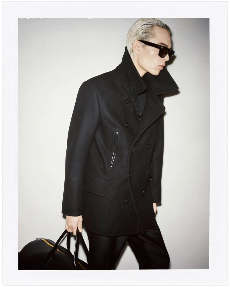 Model wearing a black coat and sunglasses holding a black duffel bag against a white seamless background