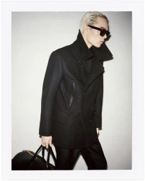 Model wearing a black coat and sunglasses holding a black duffel bag against a white seamless background