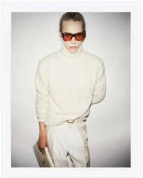 Model wearing a white turtleneck sweater, tinted sunglasses, and holding a light handbag against a white backdrop.