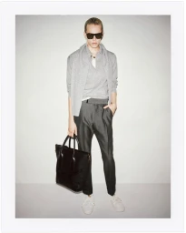 Model in grey sweater and trousers holding a black leather tote bag against a neutral studio backdrop.