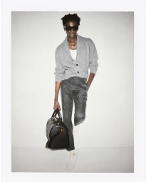 Model in grey cardigan and dark trousers holding a black duffel bag against a white studio background.