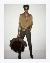 Model wearing dark sunglasses, brown shirt, scarf, and trousers while holding a black leather duffle bag against a plain wall.