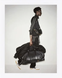 Black man in a tracksuit carrying a large duffel bag against a light studio backdrop.