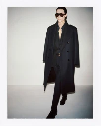 Model in dark sunglasses wearing a long black coat over a matching suit against a white studio background