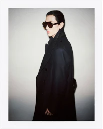 Model wearing dark sunglasses and a long black coat against a neutral gradient studio background for Tom Ford Fall 2024.