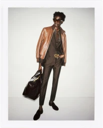 A male model wearing a brown leather jacket and suit holding a large black bag against a seamless white studio backdrop.