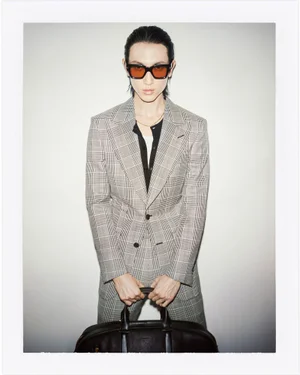 Model in a grey plaid suit wearing sunglasses and holding a dark leather bag against a white background.