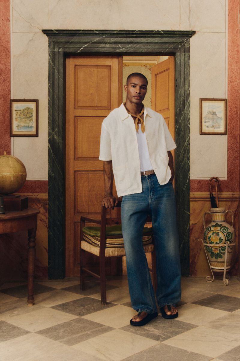 Man in white shirt and jeans leans against chair in an interior with tiled floor, decorative walls, paintings, and doorway.
