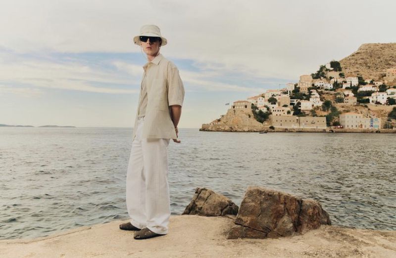 Man in casual outfit with a shirt, pants, sunglasses and bucket hat stands on a stone pier by the water with buildings in the background.