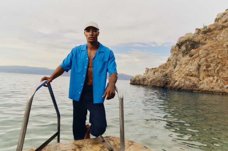 Man in unbuttoned blue shirt and dark pants stands on steps leading to ocean, with rocky coast and cloudy sky in the background.