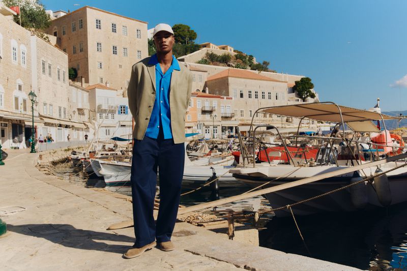 Model Niyo Malik wearing a beige blazer, bright blue shirt, and navy trousers on a sunny boat dock for Zara Man.