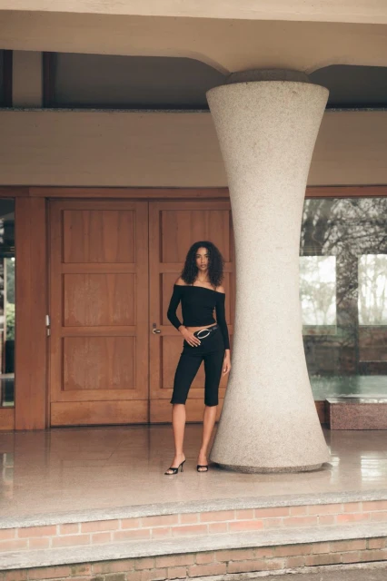 A woman in a black outfit stands in front of a building with a large column and wooden doors in a fashion-style shot.