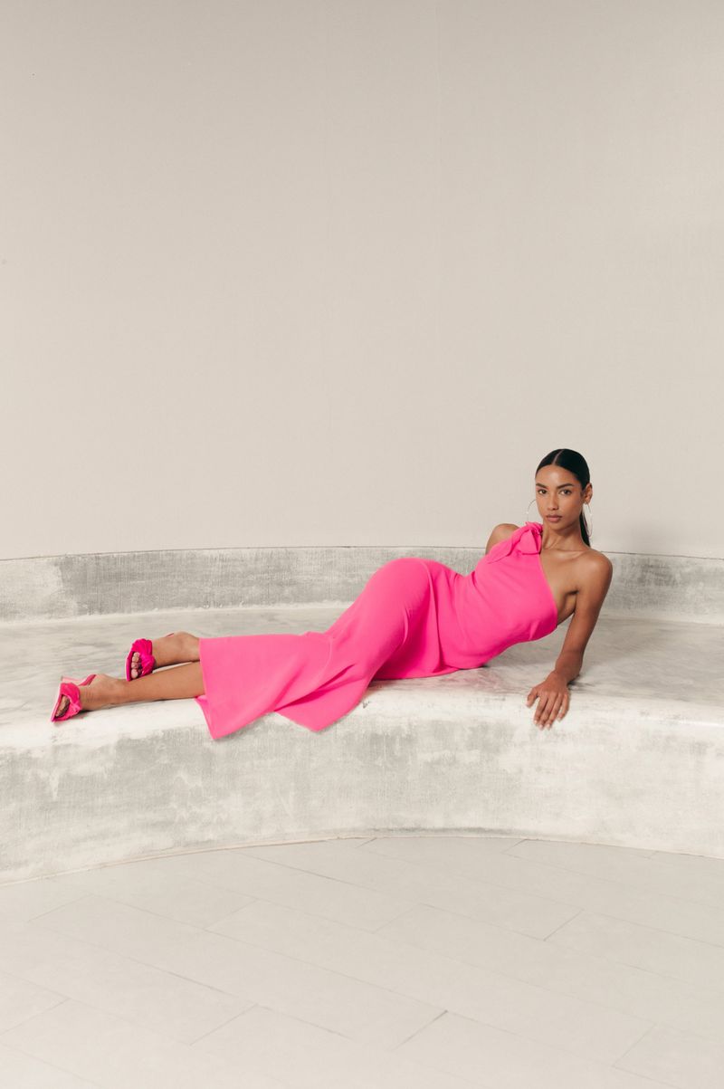 Adult female fashion model in a pink one-shoulder dress and pink heels lying on a gray concrete platform in a photo studio.