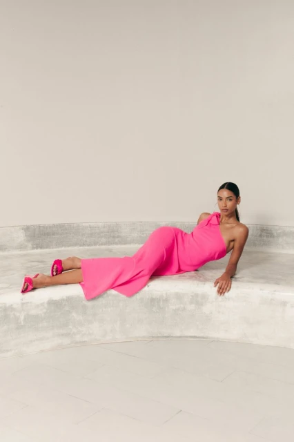 Adult female fashion model in a pink one-shoulder dress and pink heels lying on a gray concrete platform in a photo studio.