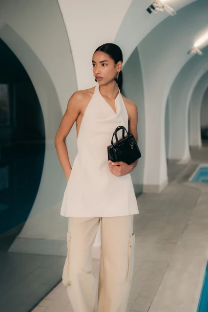 Adult woman in a cream-colored top and cargo pants holds a black handbag in a minimalist indoor setting with arched walls.