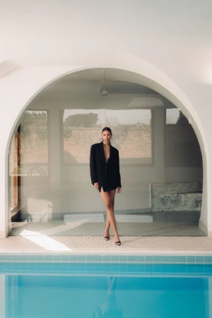 A woman in a black blazer poses near a turquoise pool, blending fashion with a serene, summery ambiance in an indoor-outdoor setting.