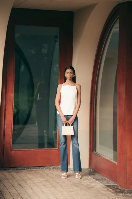 Full body shot of a woman in jeans and a white top standing in front of a glass door with a purse, posing in natural light.