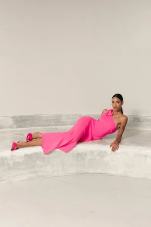 Model in vibrant pink one shoulder dress reclining on curved concrete architecture for Alessandra Milano SS24