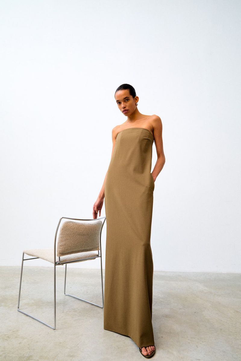 Indoor shot of an adult female model wearing a tan strapless dress standing next to a beige chair in a minimalist setting.