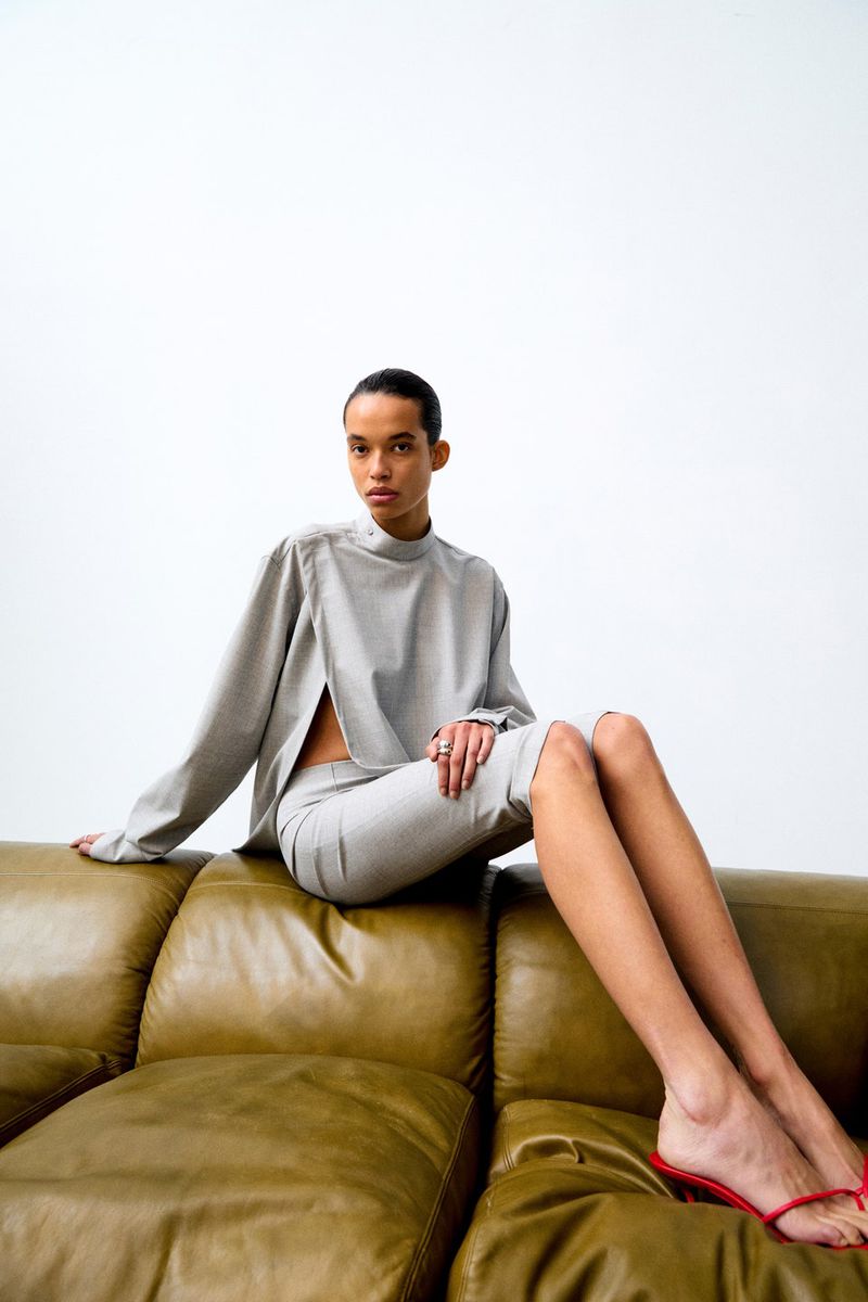 A woman with short dark hair in a grey long-sleeved top and skirt is sitting on an olive-green leather sofa in a light studio.
