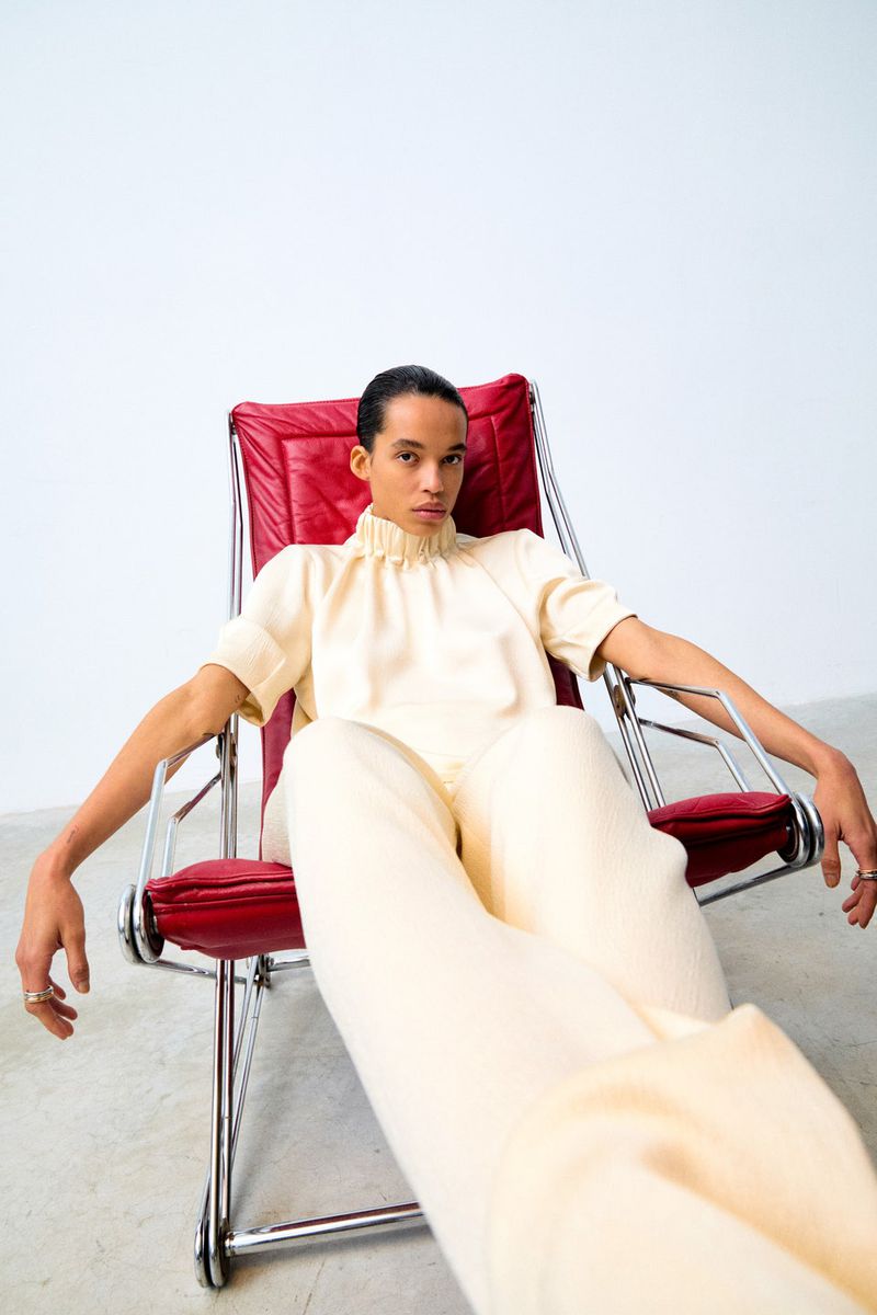 Person sits in a red lounge chair, facing the camera, wearing a beige high-necked short sleeve shirt and wide-legged pants against a white wall.