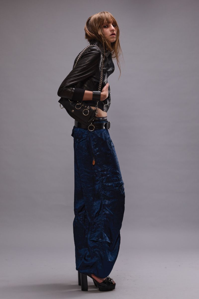 Full-length studio shot of a young adult female fashion model posing in a black leather jacket and blue patterned trousers with platform heels.