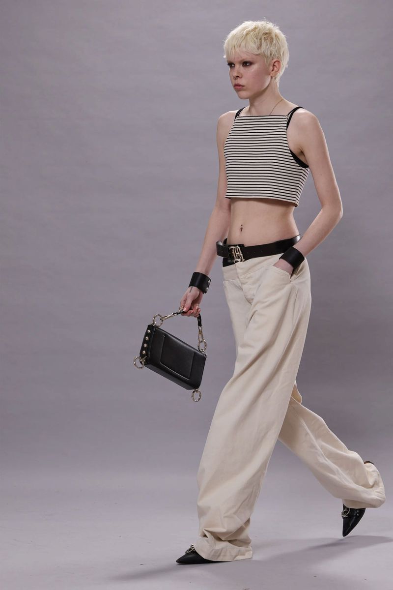Fashionable woman with platinum blonde hair walks in a striped crop top, baggy pants, and black accessories against a grey backdrop.