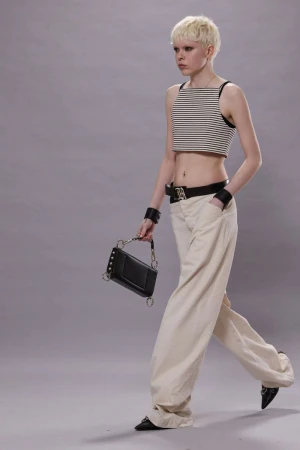 Fashionable woman with platinum blonde hair walks in a striped crop top, baggy pants, and black accessories against a grey backdrop.