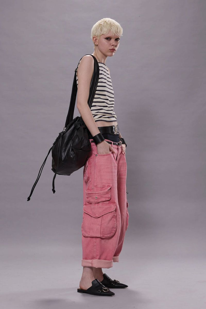 Adult female with short blonde hair posing in front of a grey background, wearing a striped shirt, pink cargo pants, and black flats.