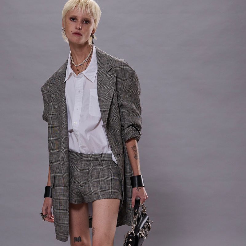 A slender fashion model with short blonde hair poses in a studio, wearing an oversized plaid blazer with matching shorts and a white shirt.