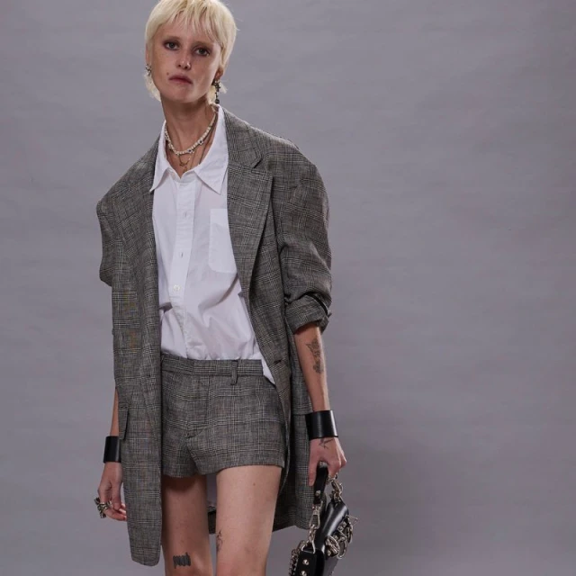 A slender fashion model with short blonde hair poses in a studio, wearing an oversized plaid blazer with matching shorts and a white shirt.