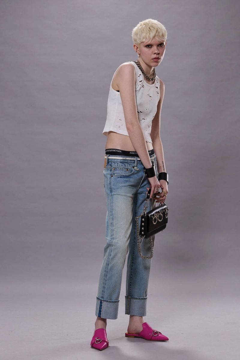Full-length studio shot of a slender person with short blonde hair wearing a white tank top, jeans, and pink mules with a black purse.