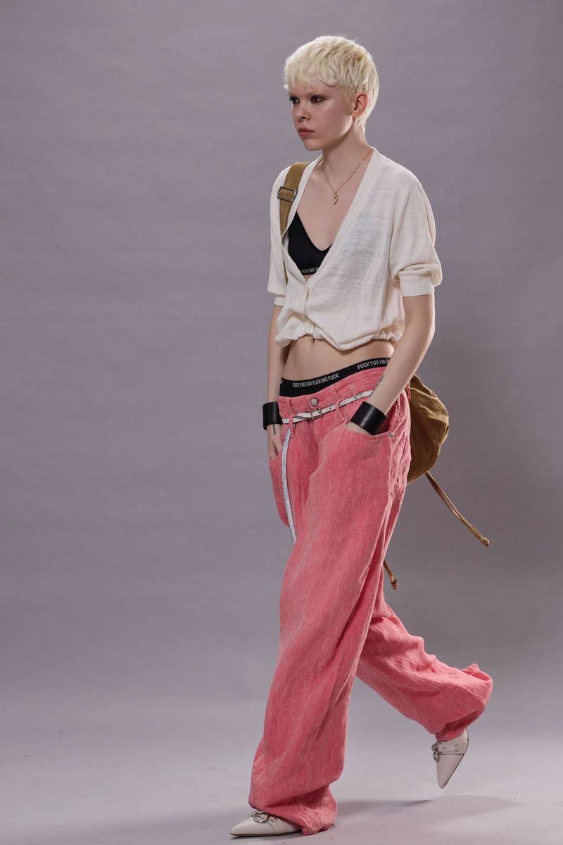 A fashionably dressed young adult woman is walking in pink pants and a cardigan, accented by a cross necklace and shoulder bag, against a plain backdrop.
