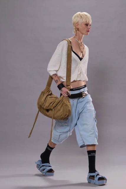 Fashionable adult woman walks in a photo studio wearing a white cropped cardigan, denim shorts, platform sandals, and a brown shoulder bag.