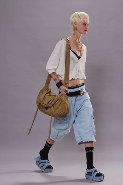 Woman with blonde hair walks in light blue cargo shorts, a cropped cardigan, black socks, and sandals with a light brown shoulder bag.