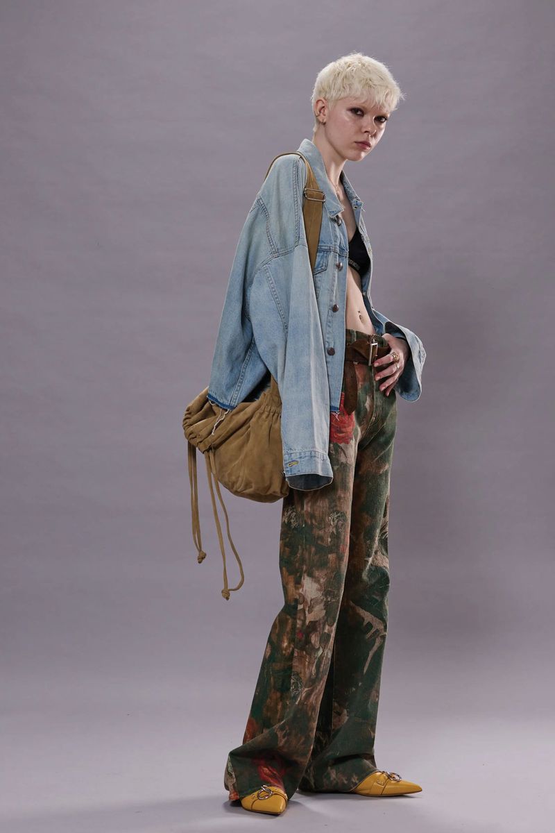 Full shot of a young adult woman posing in a light-blue denim jacket, artfully patterned jeans, and low yellow heels against a grey backdrop.