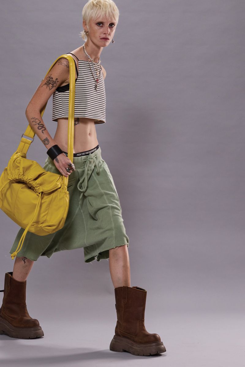 A slender woman with short blonde hair poses in a studio wearing a striped crop top, green skirt, brown boots, and a yellow shoulder bag.